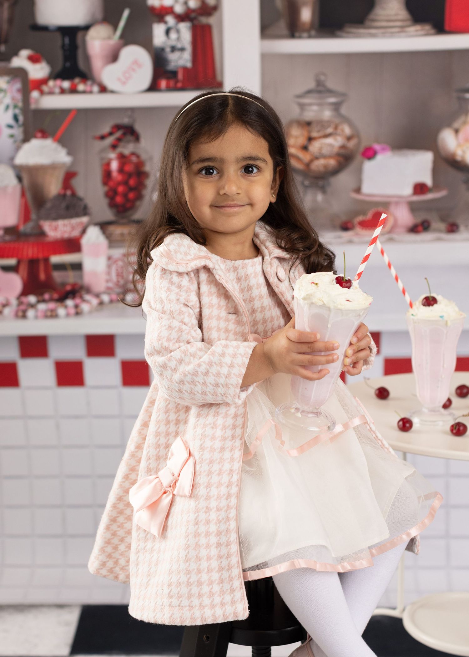 A child in a pink polka dot coat poses with a white cake in a retro-styled kitchen setting with red accents.