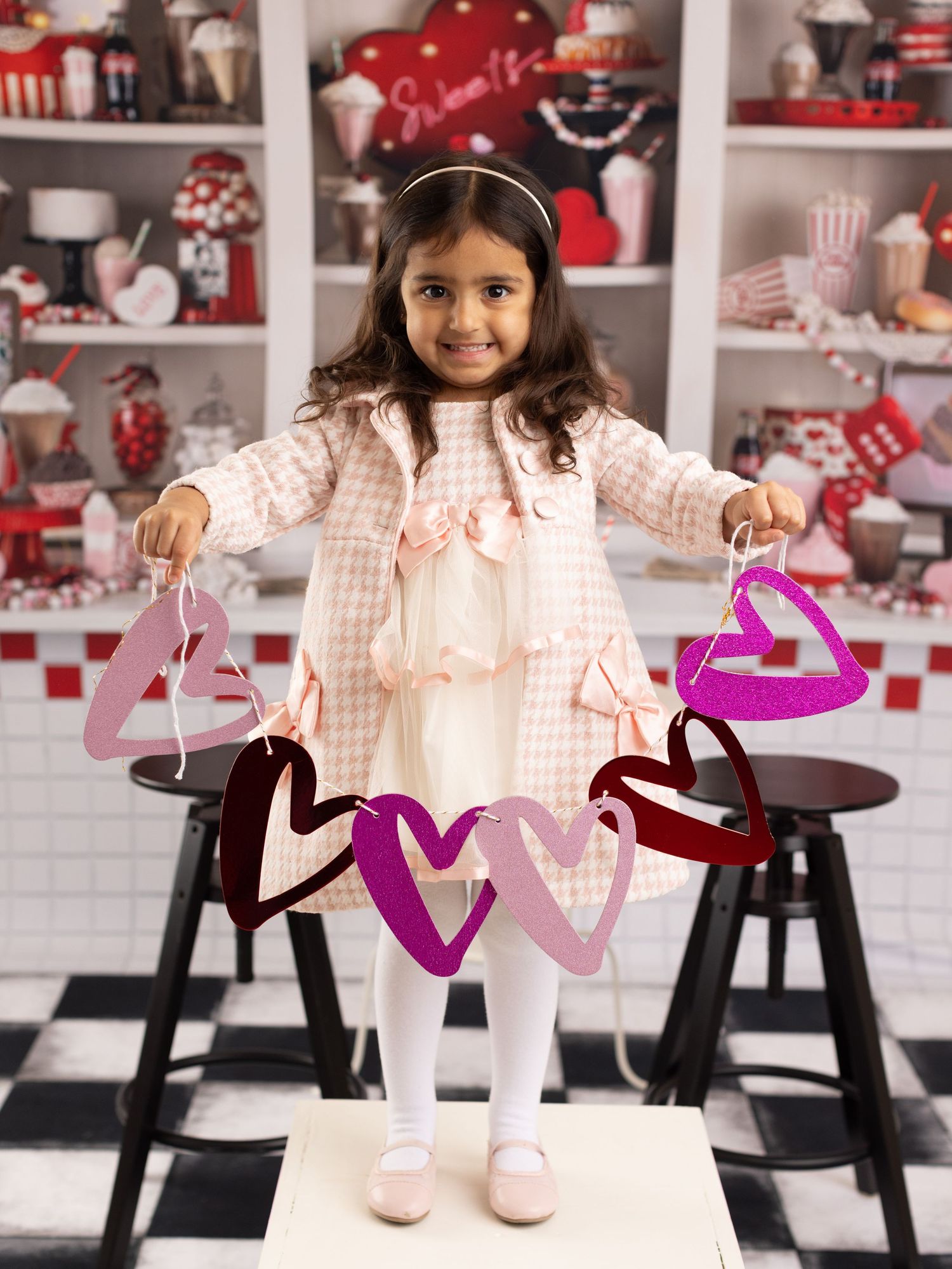 A young girl in a pink gingham dress poses in a red and white retro-styled diner setting.