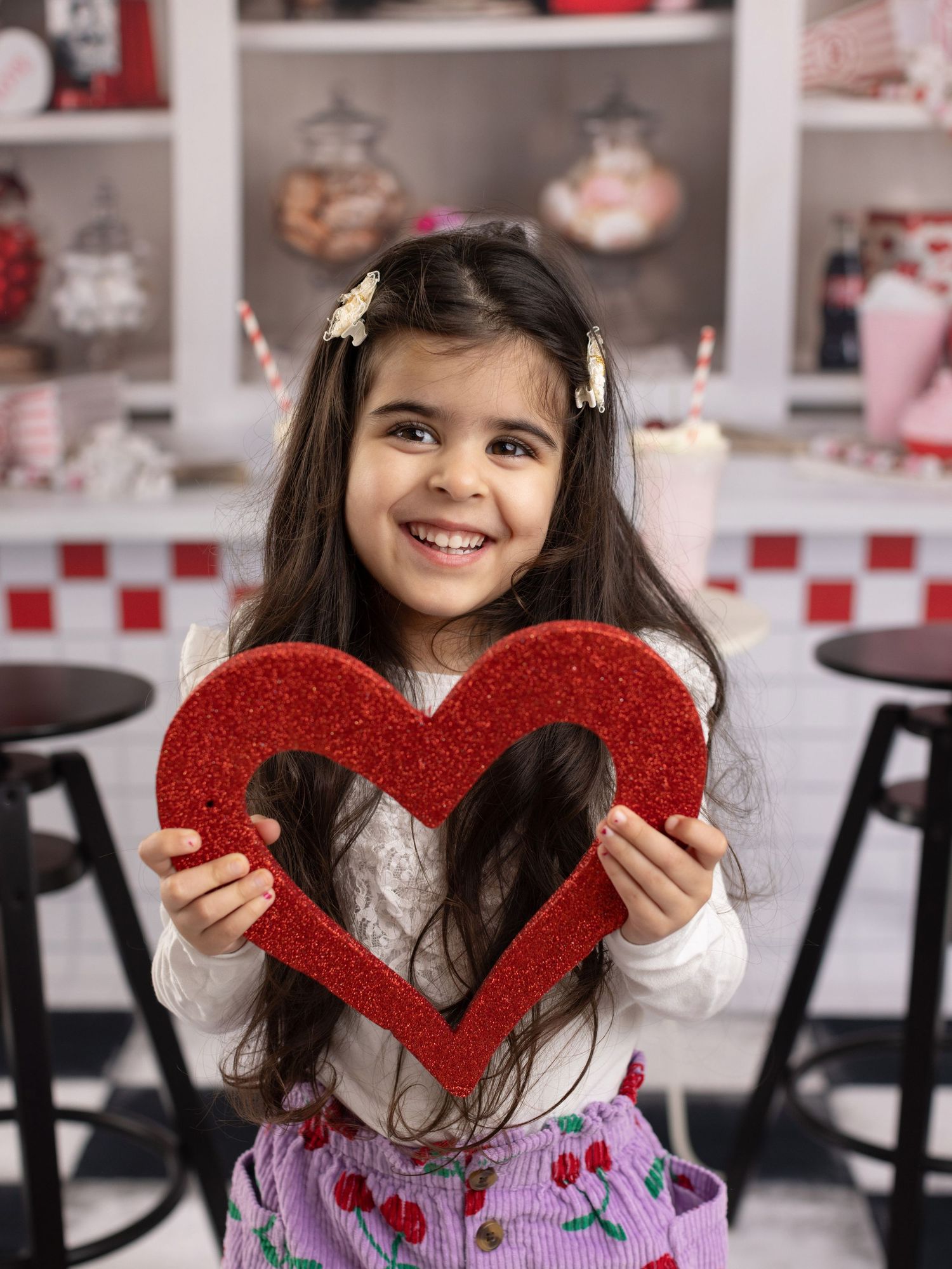 A young child in a white sweater holds a sparkly red glitter heart decoration in a diner-style setting.