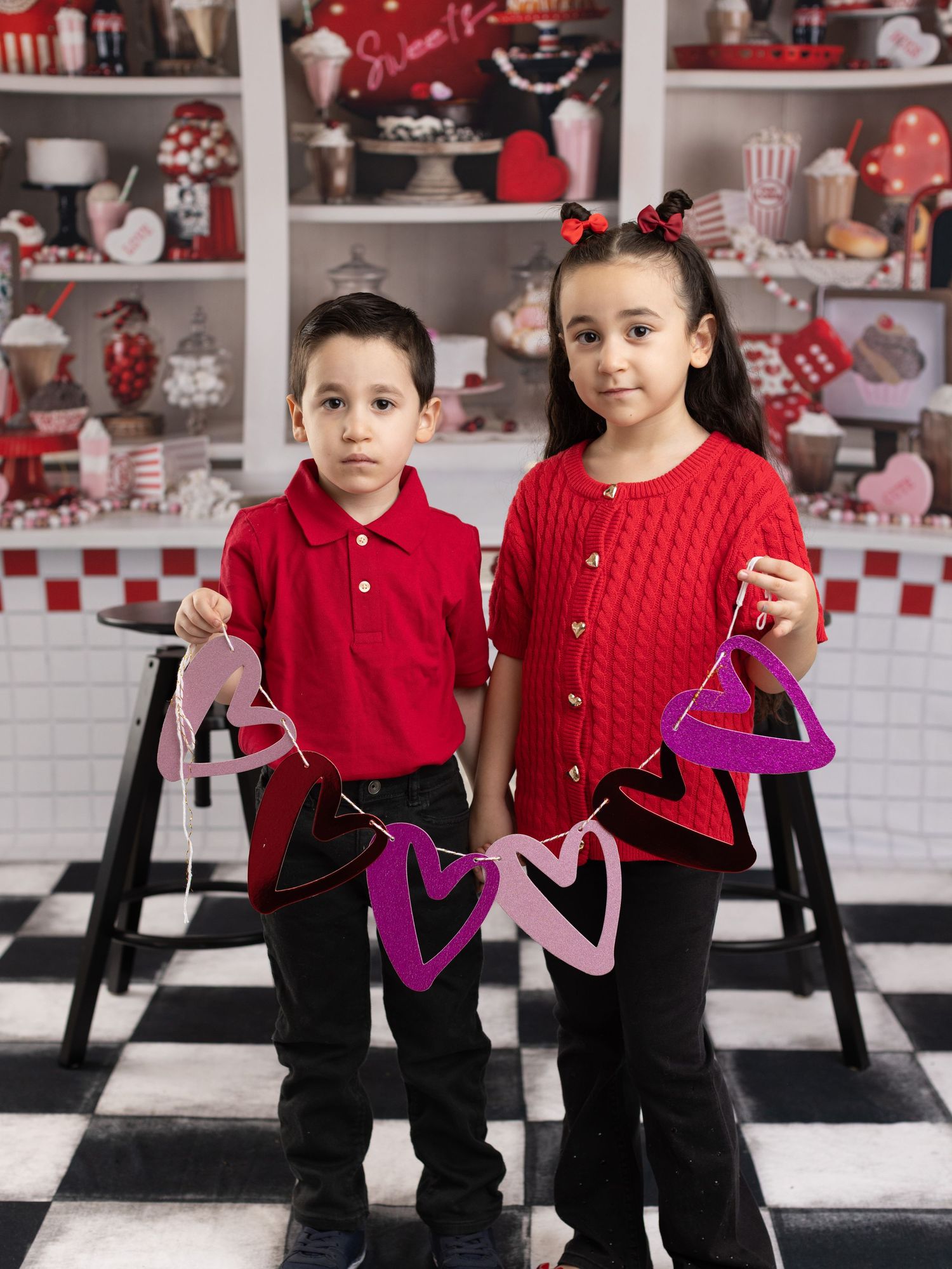 Two children in matching red sweaters pose playfully in a retro diner-themed setting with black and white checkered floor.