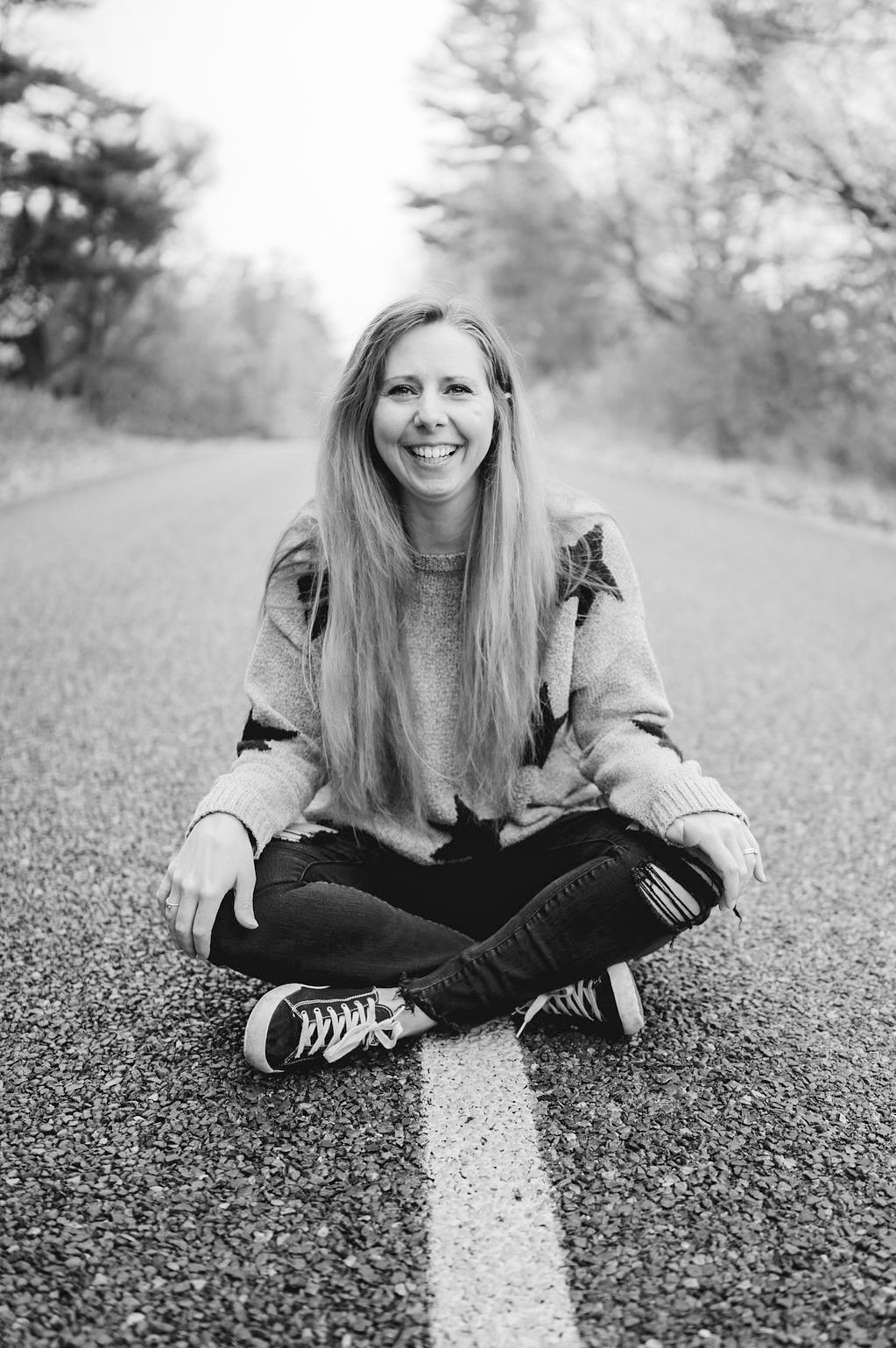 A person in a grey sweater sits on an autumn road with yellow line markings during fall season.