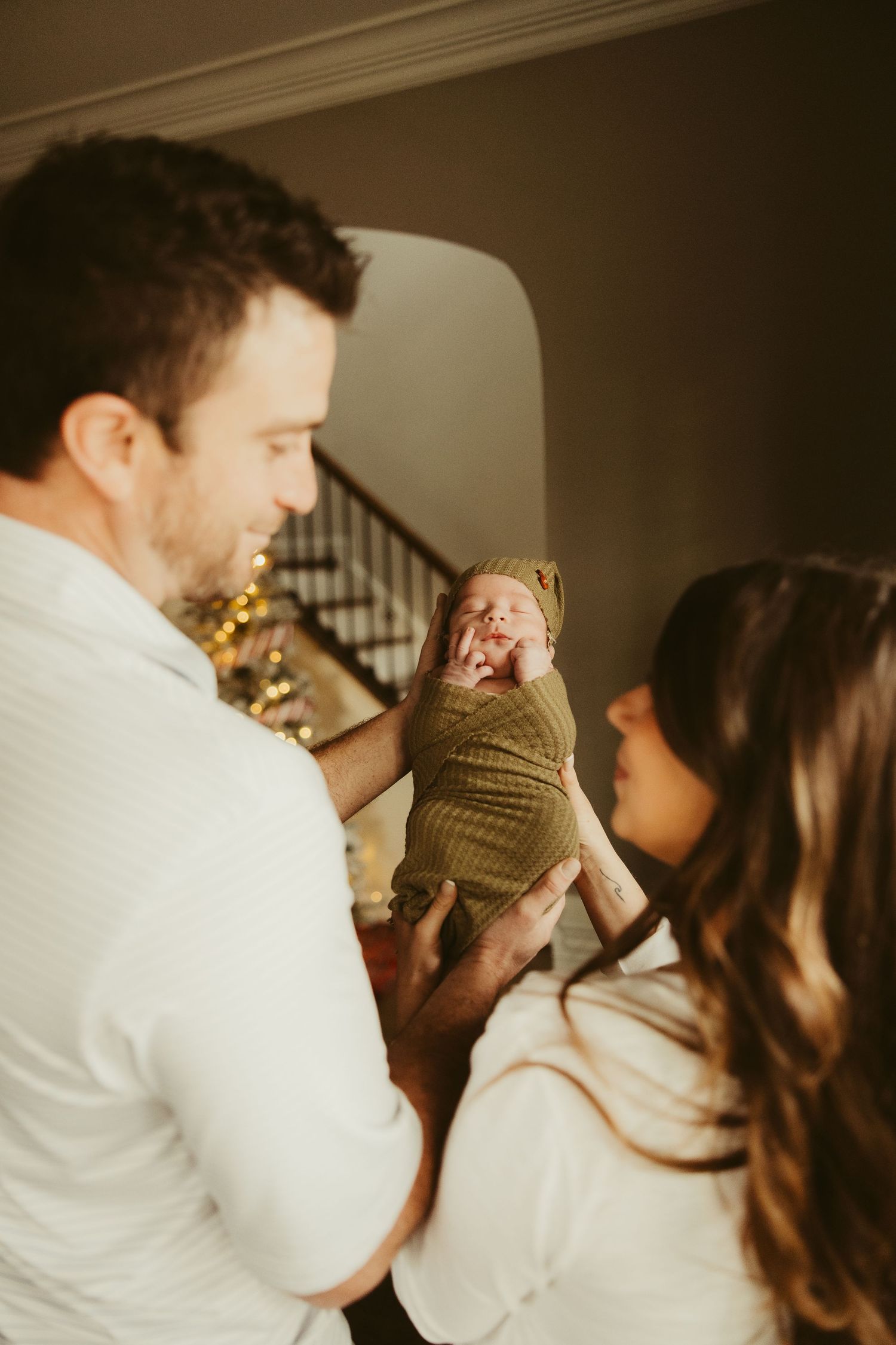 New parents smiling at swaddled newborn in olive wrap during cozy home photo session.