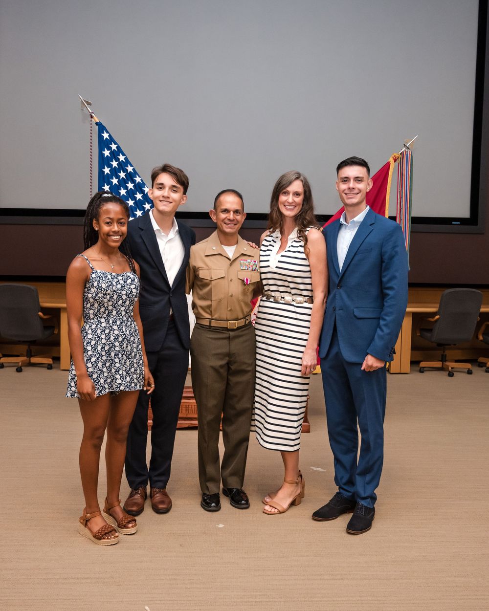 A group stands together in front of an American flag and projection screen in a formal setting.