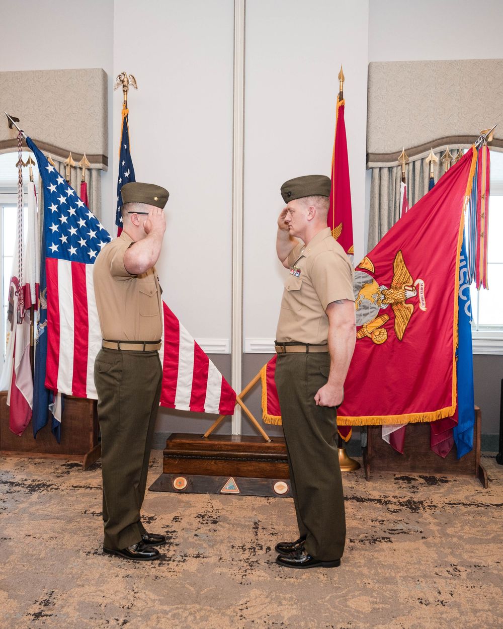 Military personnel in uniform perform a ceremonial flag presentation in front of a display of American and service flags.