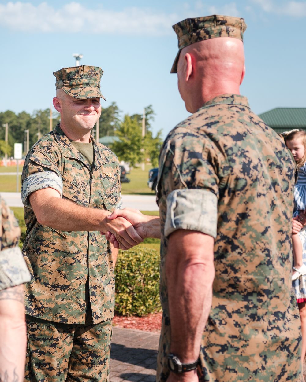 Military service members in camouflage uniforms interact during an outdoor ceremony on a sunny day.