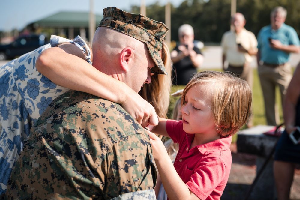 A service member in camouflage uniform shares an emotional embrace with a young child wearing a pink shirt.