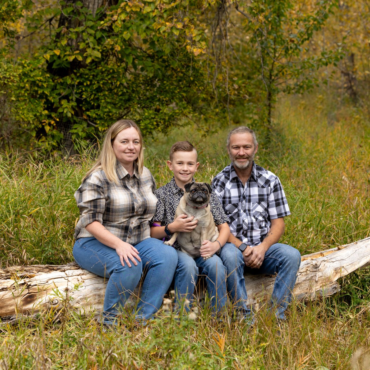 A family sits together on a fallen log in an autumn woodland setting with their pet dog.