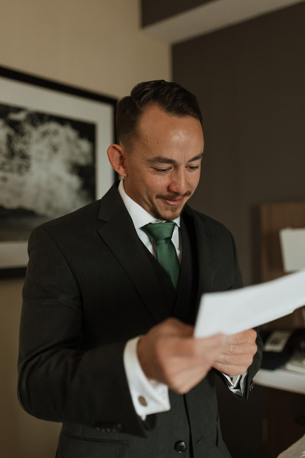 A groom in a dark suit and green tie reads a letter from his bride while standing in a room.