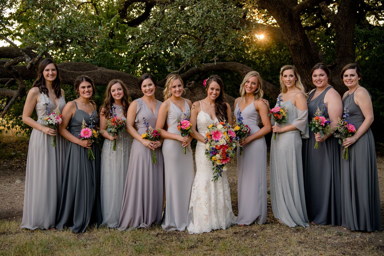A bridal party in flowing gray and slate blue dresses poses together outdoors during a sunset wedding ceremony.