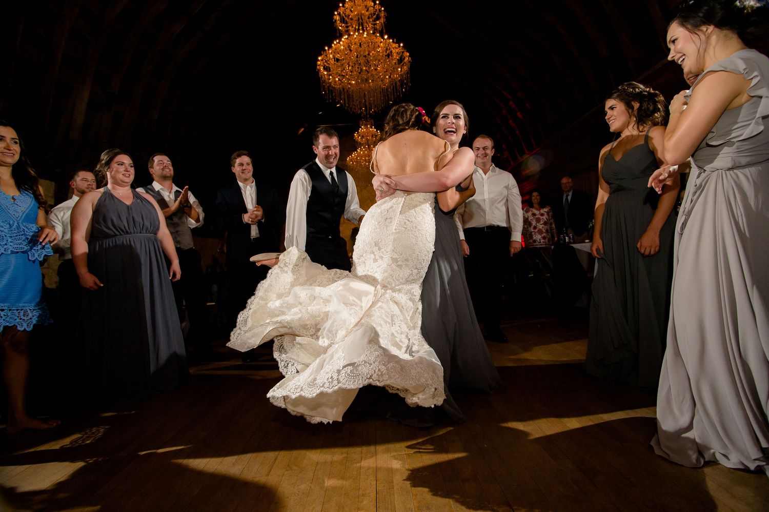 A wedding party celebrates on a dance floor with bridesmaids in gray dresses and a bride in a flowing white gown.