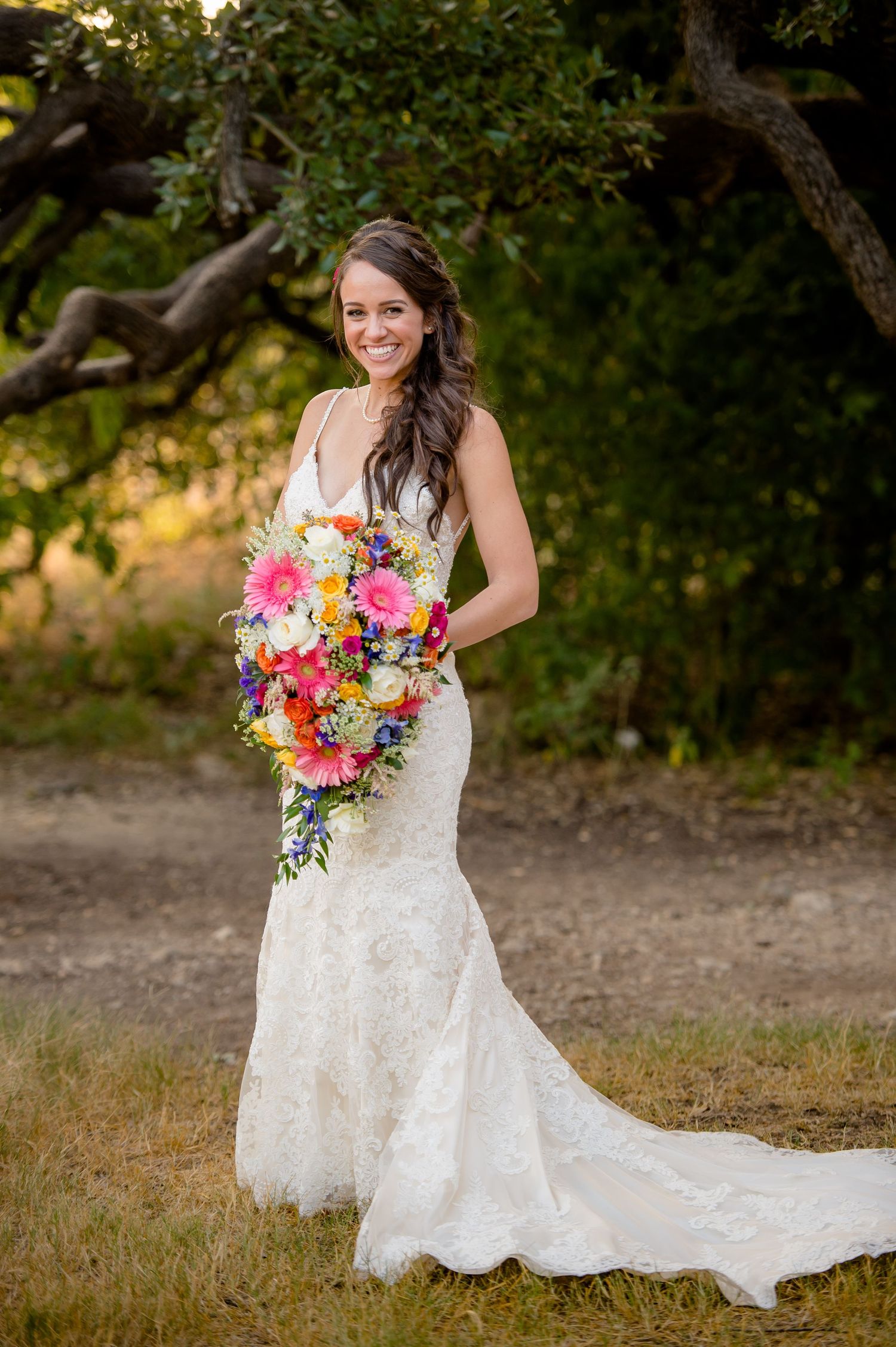 A bride in a white lace mermaid wedding dress holds a vibrant pink and orange bouquet in an outdoor garden setting.