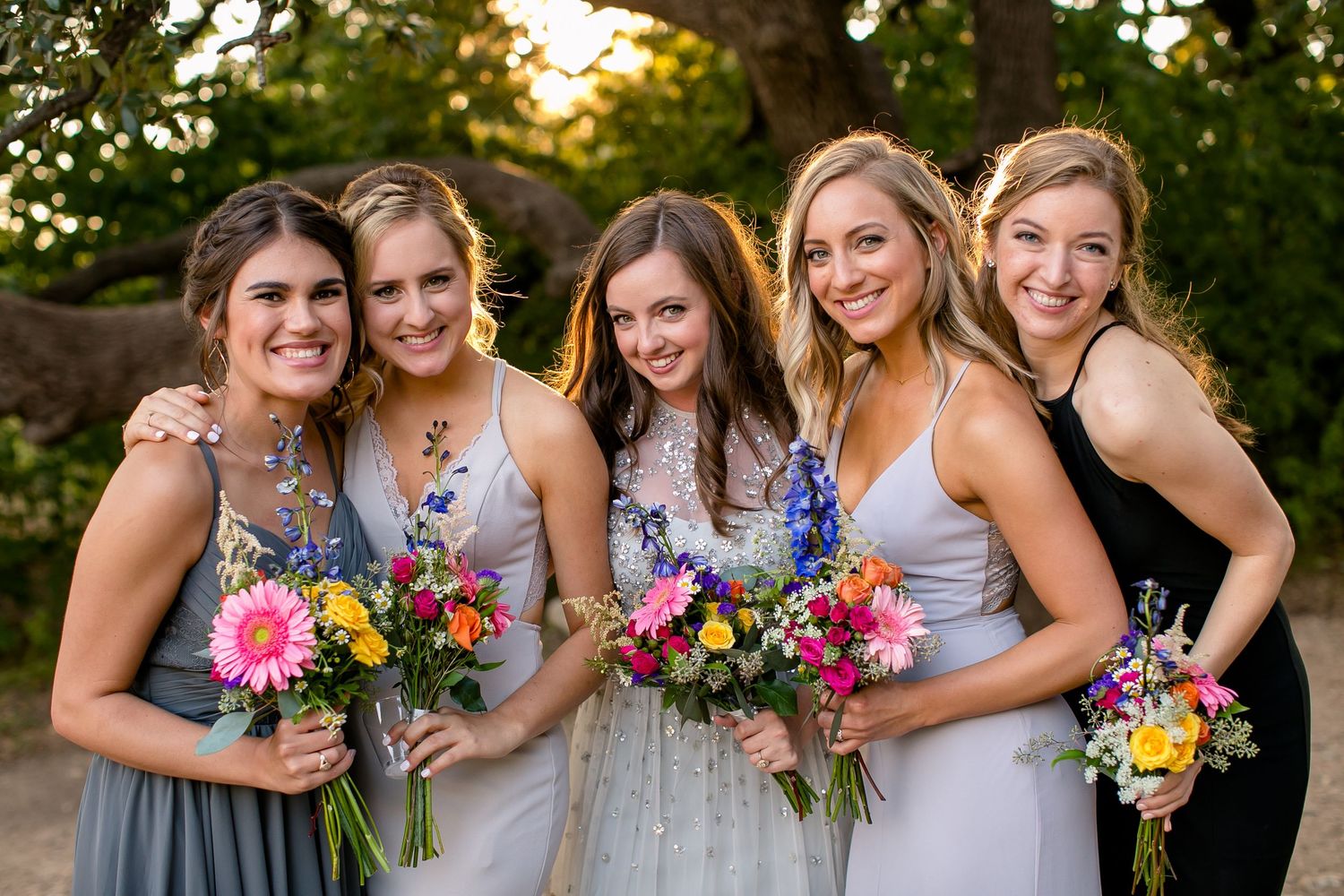 Bridesmaids in gray dresses hold colorful summer bouquets while posing together outdoors at sunset.
