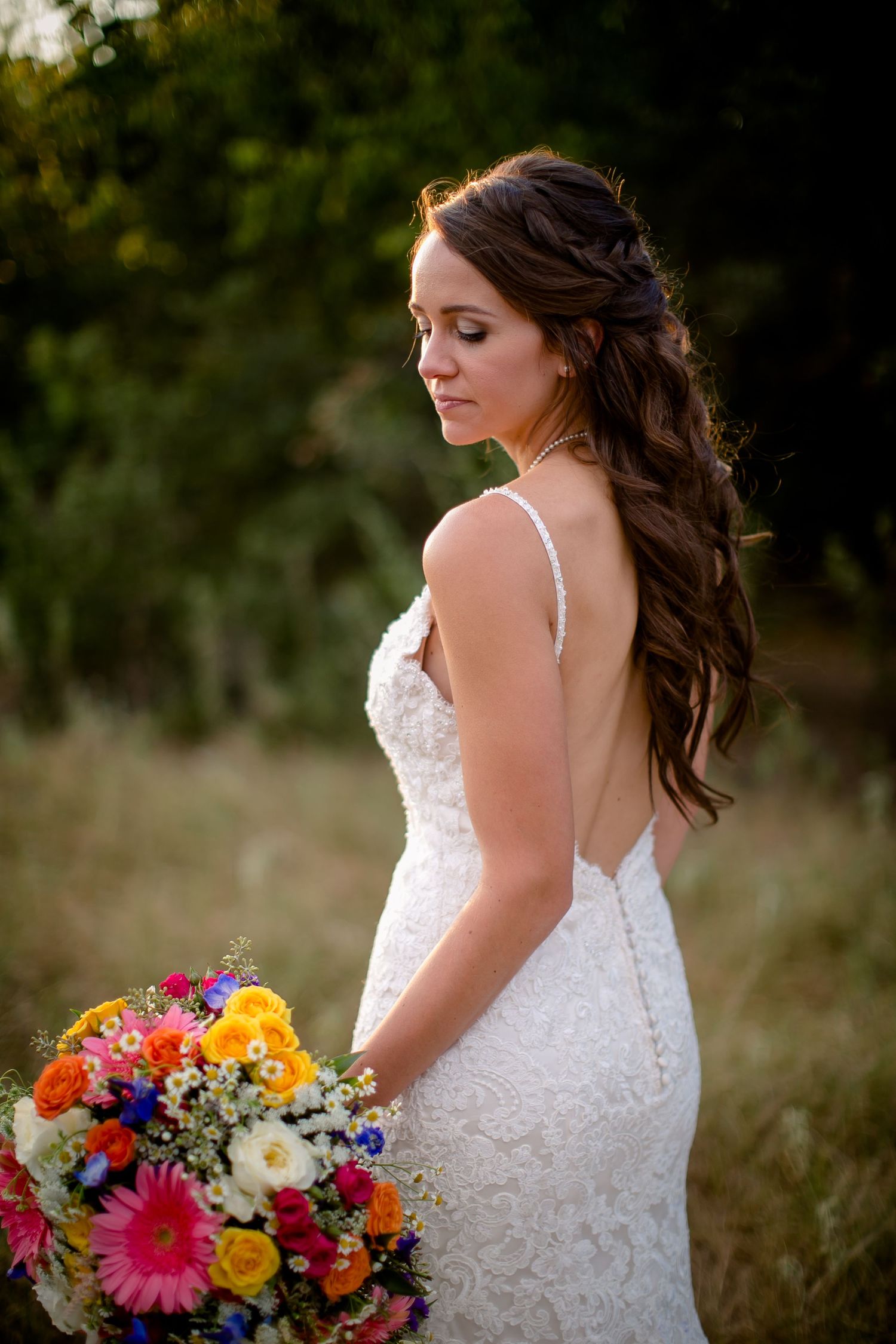 A bride in a white lace dress holds a vibrant bouquet of gerbera daisies, sunflowers and wildflowers in an outdoor setting.