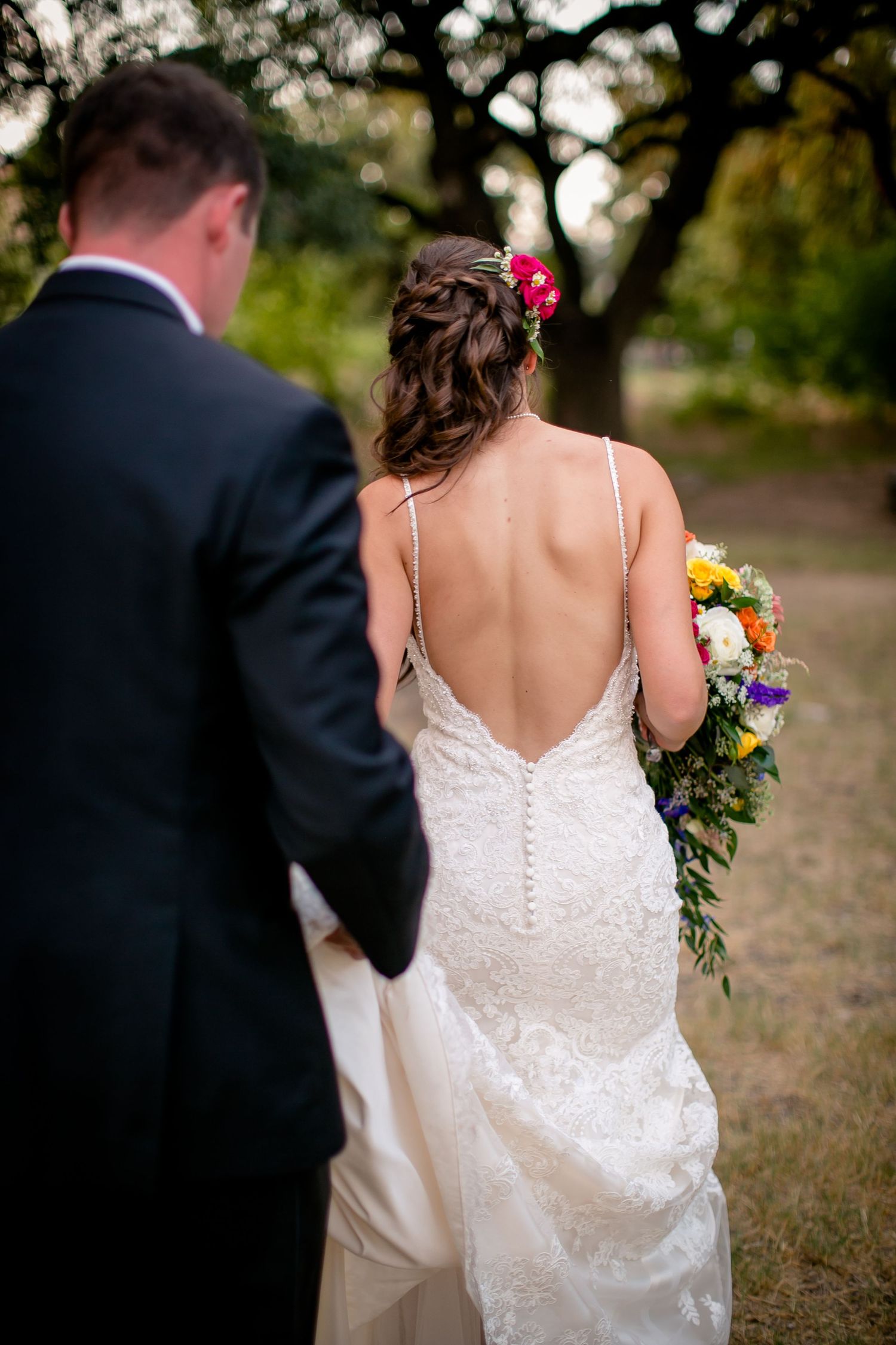 A bride in a white lace dress with spaghetti straps holds a colorful bouquet while walking outdoors.
