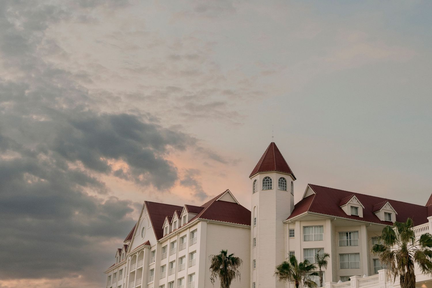 Grand white hotel building with red pitched roofs, tower, and palm trees under a dramatic cloudy sunset sky.