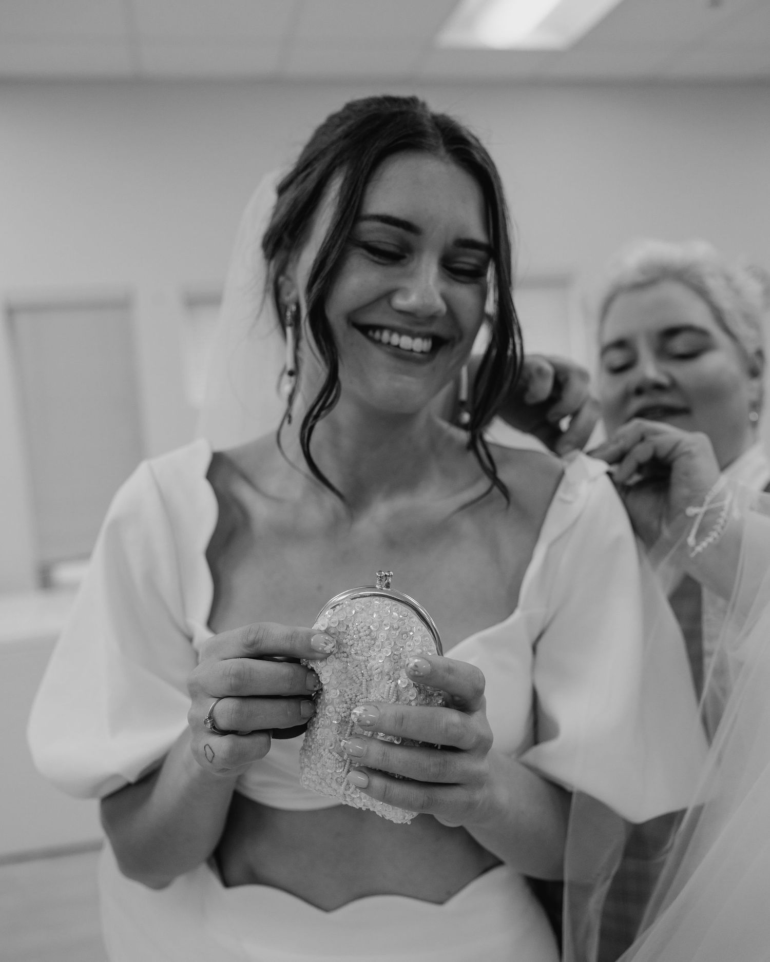 Black and white photo of smiling bride holding a beaded clutch while getting ready with bridesmaid.