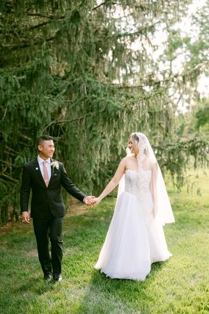 A bride and groom walk hand in hand on green grass beneath tall evergreen trees on their wedding day.