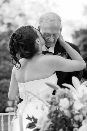 Black and white photo of a bride kissing an elderly man in a tuxedo at an outdoor wedding ceremony.