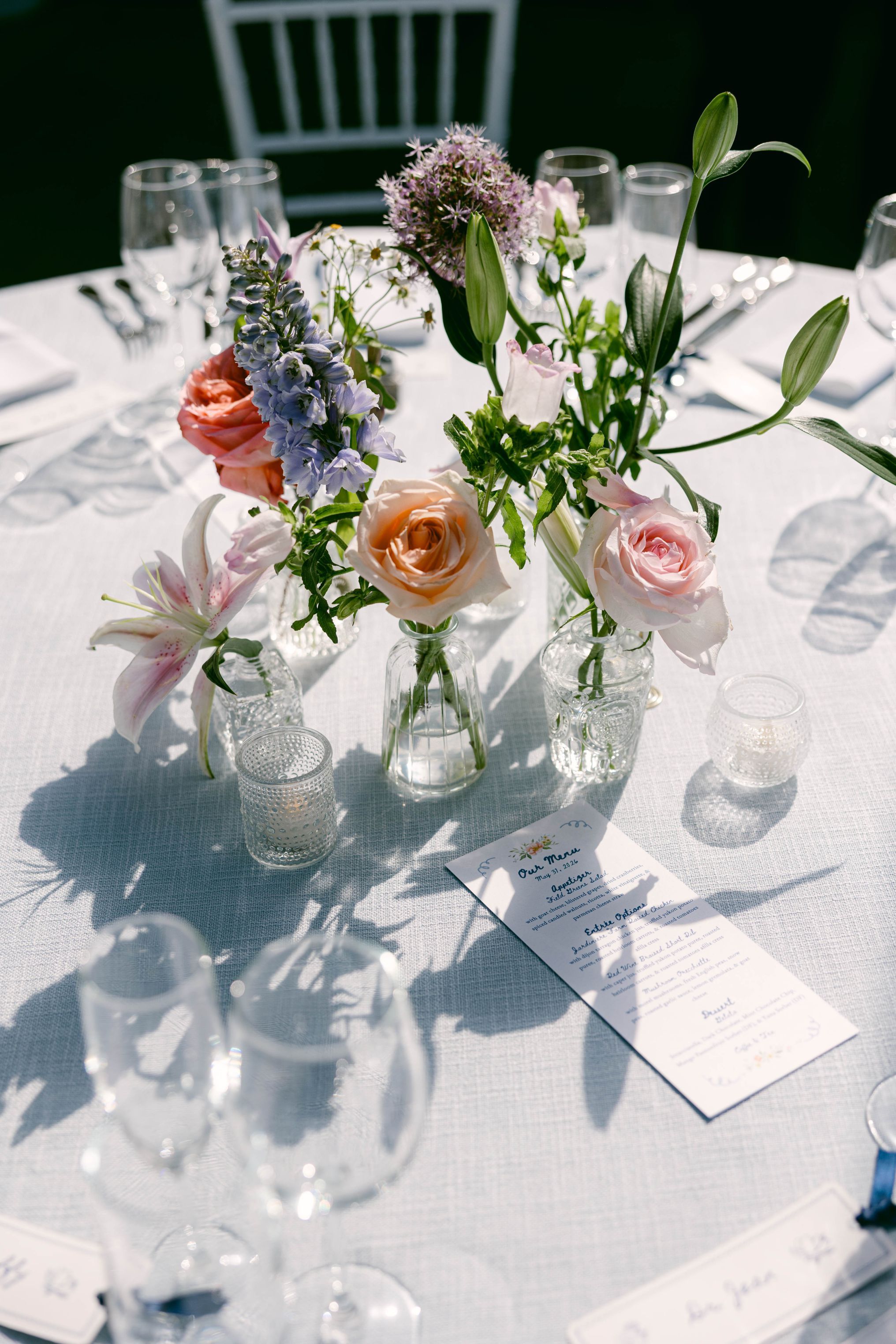 Elegant wedding table with glass bud vases holding colorful roses and wildflowers on a white linen tablecloth.