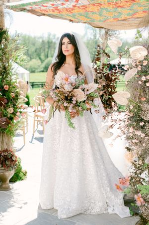 A bride in a lace wedding gown stands beneath a floral arch decorated with coral and peach blooms.