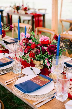 Elegant wedding table setting with red and pink floral centerpiece, blue candles, and navy napkins on white charger plates.