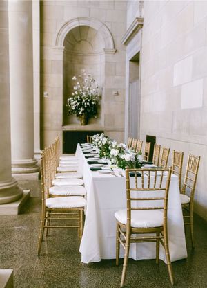 Elegant wedding reception setup with white linens and gold chiavari chairs in a grand columned hallway.
