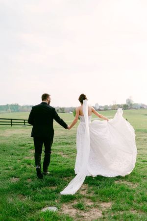Wedding couple running playfully through a green field, with the bride's dress and veil flowing in the wind.