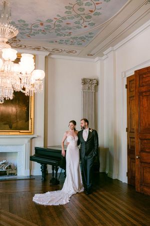 Elegant wedding couple poses in a grand historic room with ornate ceiling details and crystal chandeliers.