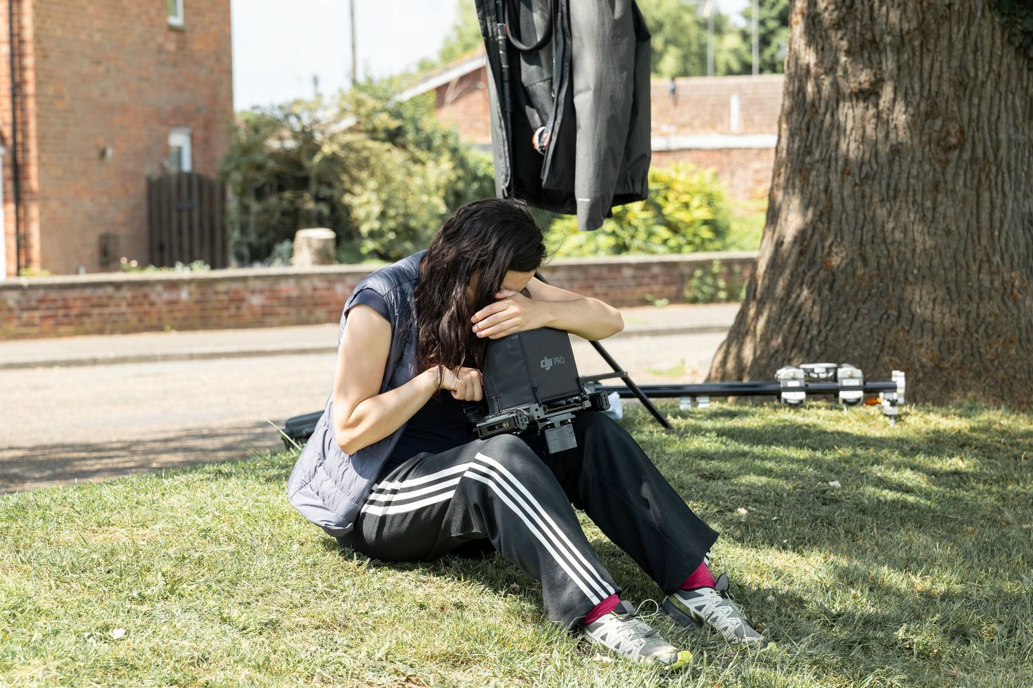 A person sits under a shady tree on a grassy lawn near brick buildings on a sunny day.