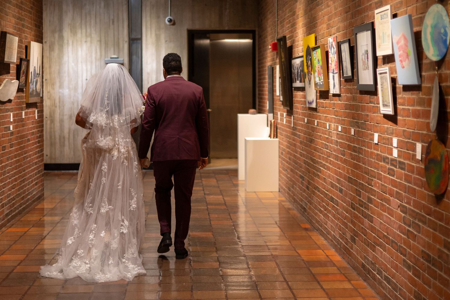 A bride and groom walk down a brick-walled hallway with artwork displayed on the walls.