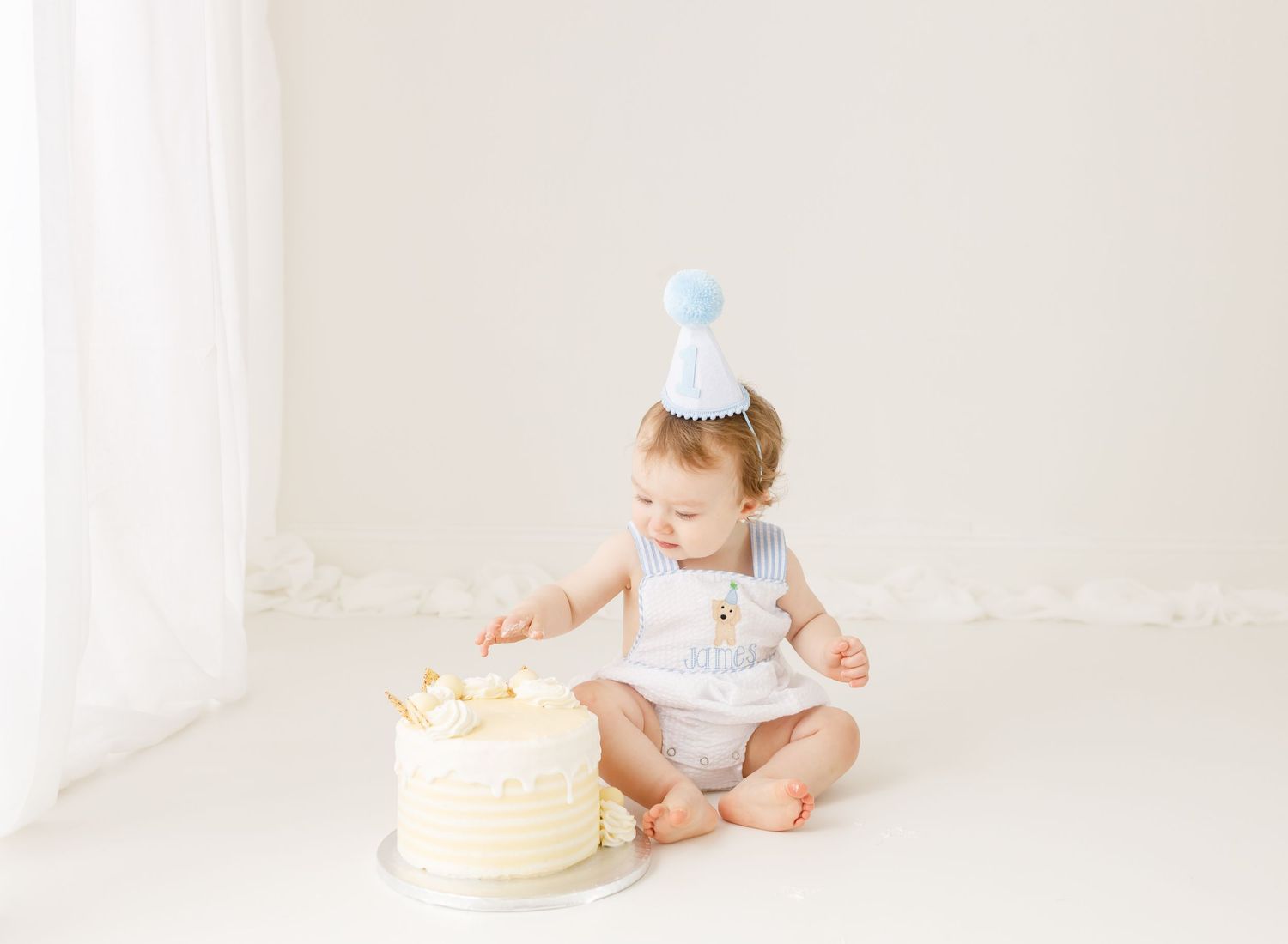 Baby in party hat sitting next to birthday cake in minimalist white setting.