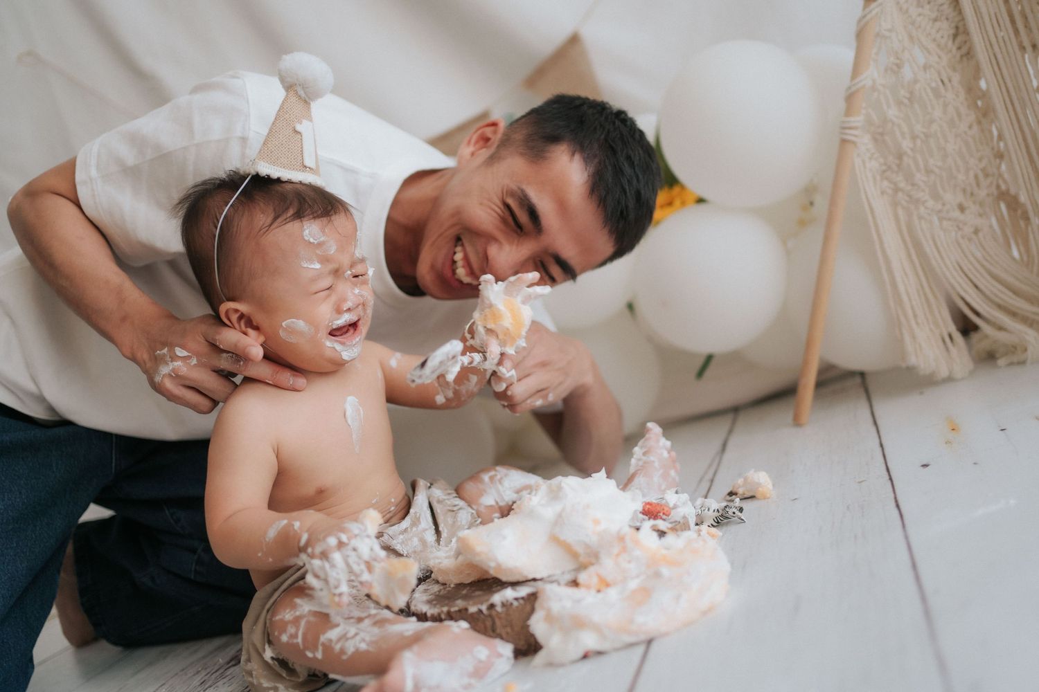 A baby celebrates their first birthday by smashing and playing with white frosted cake during a cake smash photo session.