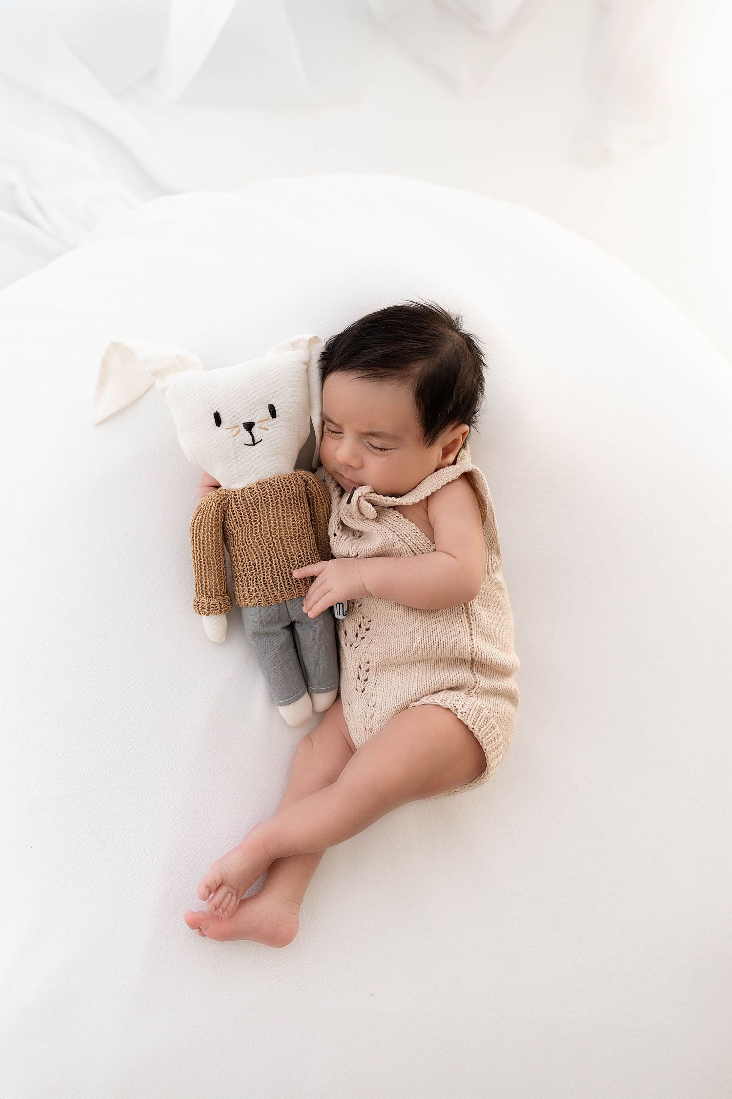 Baby in knit romper lying on white surface cuddling with plush teddy bear toy.