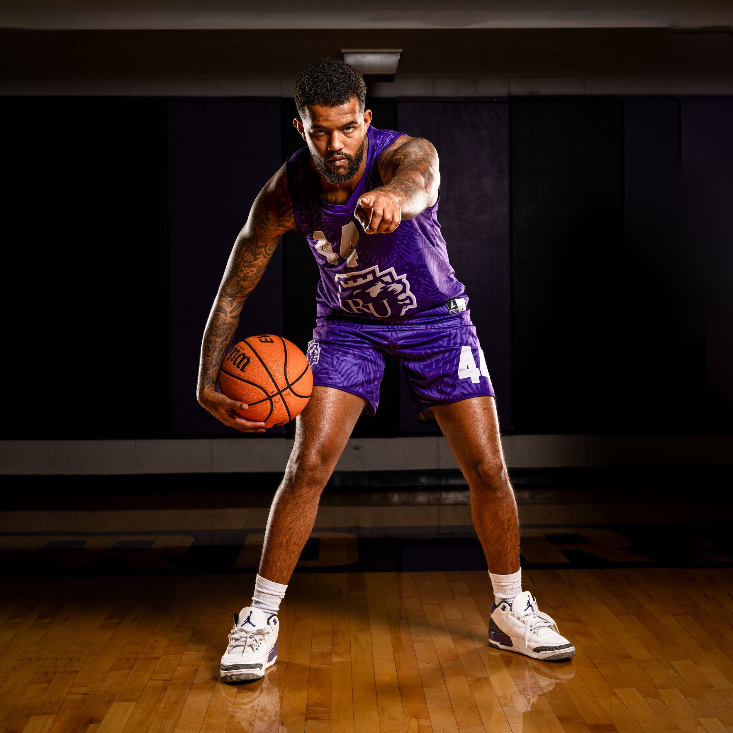 Basketball player in purple jersey crouching low with ball on wooden court during dramatic studio photoshoot.