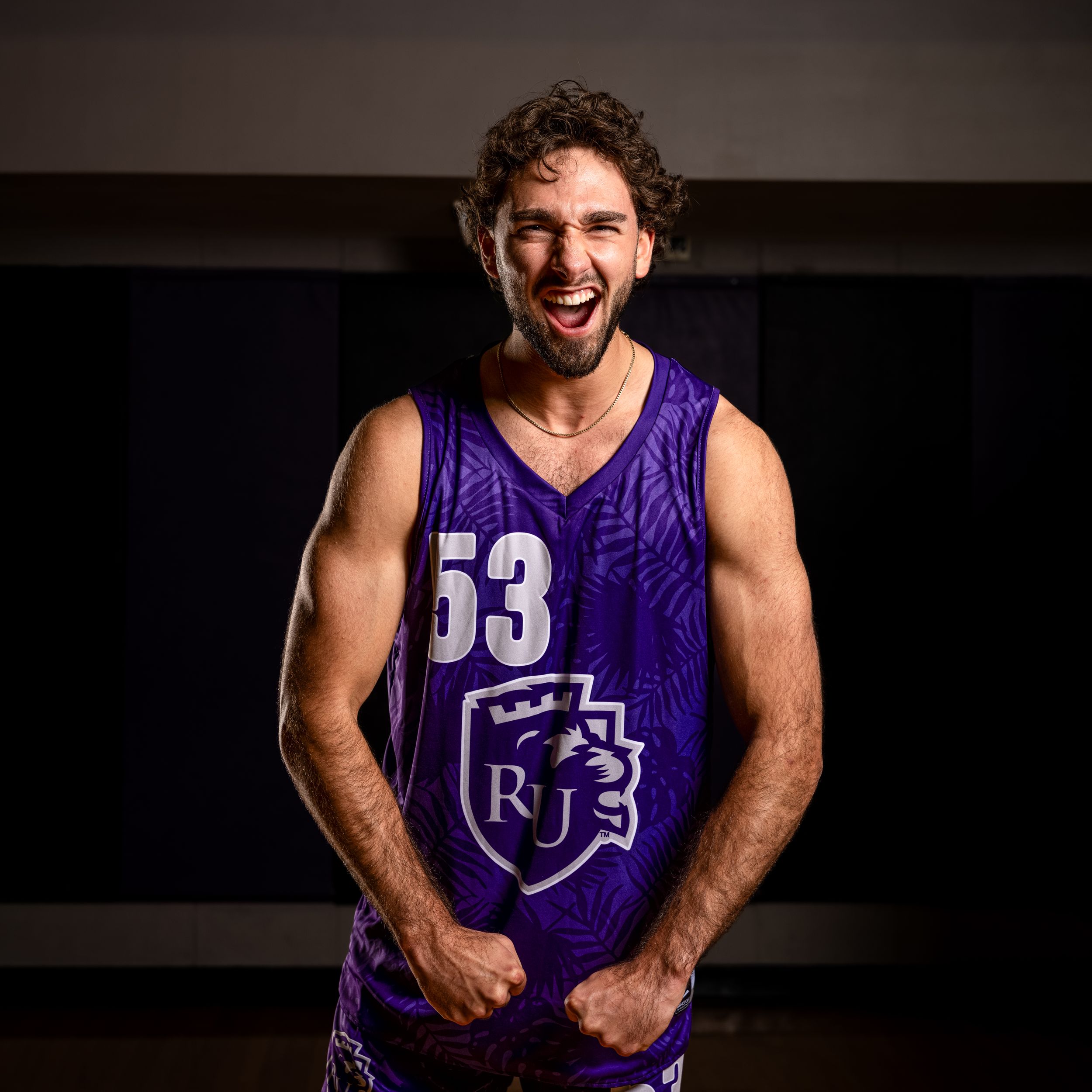Basketball player in purple jersey number 53 smiles while posing for promotional photo against dark background.