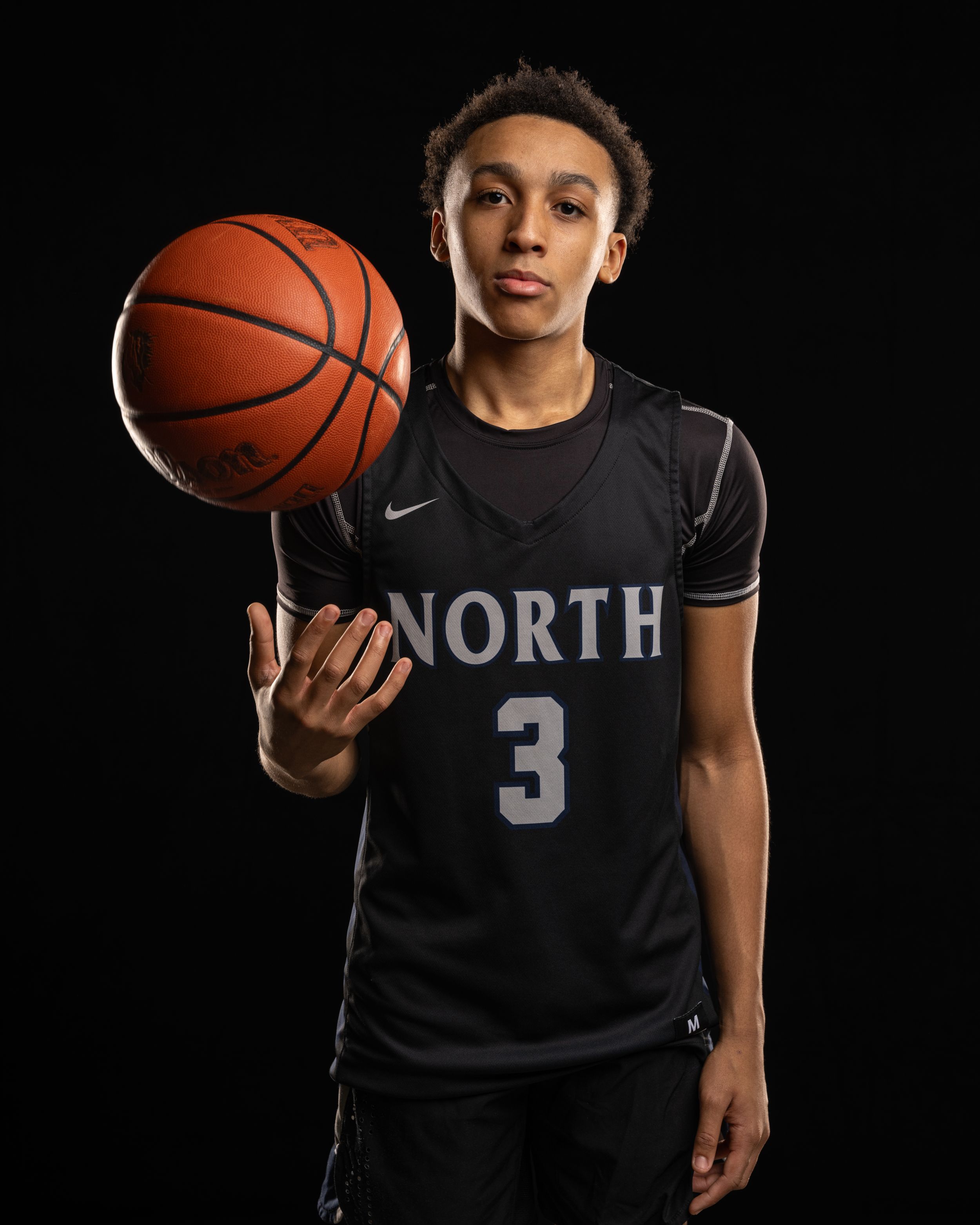 Basketball player in black North jersey number 3 poses with ball against dark background in professional portrait.