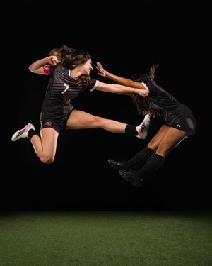 Two female soccer players in black uniforms leap dynamically against a dark background on a green field.