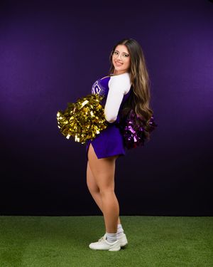 Cheerleader in a navy uniform with gold pom-poms performs a low kick pose against a purple backdrop.