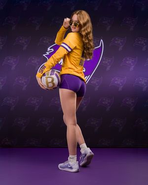 LSU cheerleader in gold uniform performs dynamic dance move against purple backdrop with repeated logo pattern.