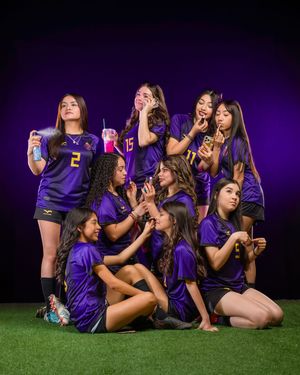 A group in matching purple jerseys pose playfully with drinks on artificial turf against a dark purple background.