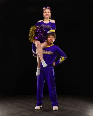 Two cheerleaders in purple uniforms performing a stunt with flyer holding gold pom-poms overhead.