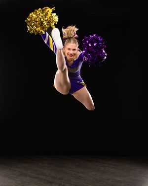 Cheerleader in purple uniform jumps mid-air holding gold and purple pom-poms against black background.