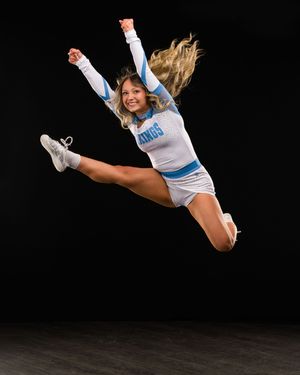 Cheerleader in blue and white uniform mid-jump against black background, hair flowing, smiling with legs extended.