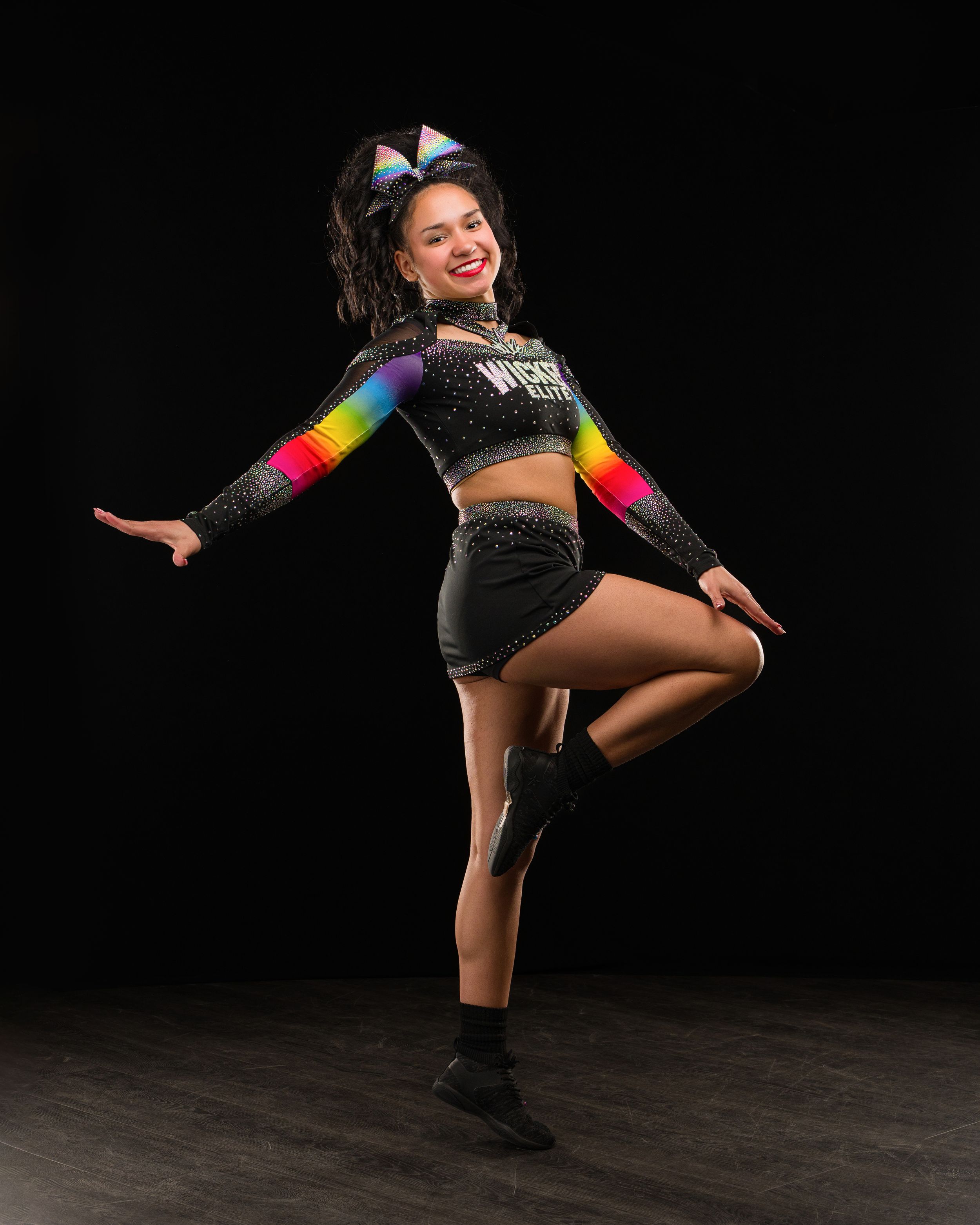 Cheerleader in rainbow and black uniform poses dynamically against dark background, wearing colorful bow and sparkly crop top.