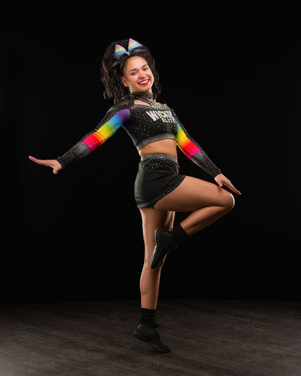 Cheerleader in rainbow and black uniform poses dynamically against dark background, wearing colorful bow and sparkly crop top.