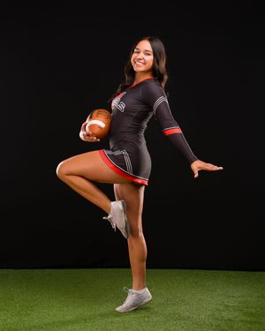 A cheerleader in a black uniform performs a high kick routine while holding a football on a turf field.
