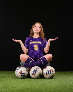 A young female soccer player in purple number 8 jersey sits cross-legged on soccer balls with arms raised in a shrug pose.