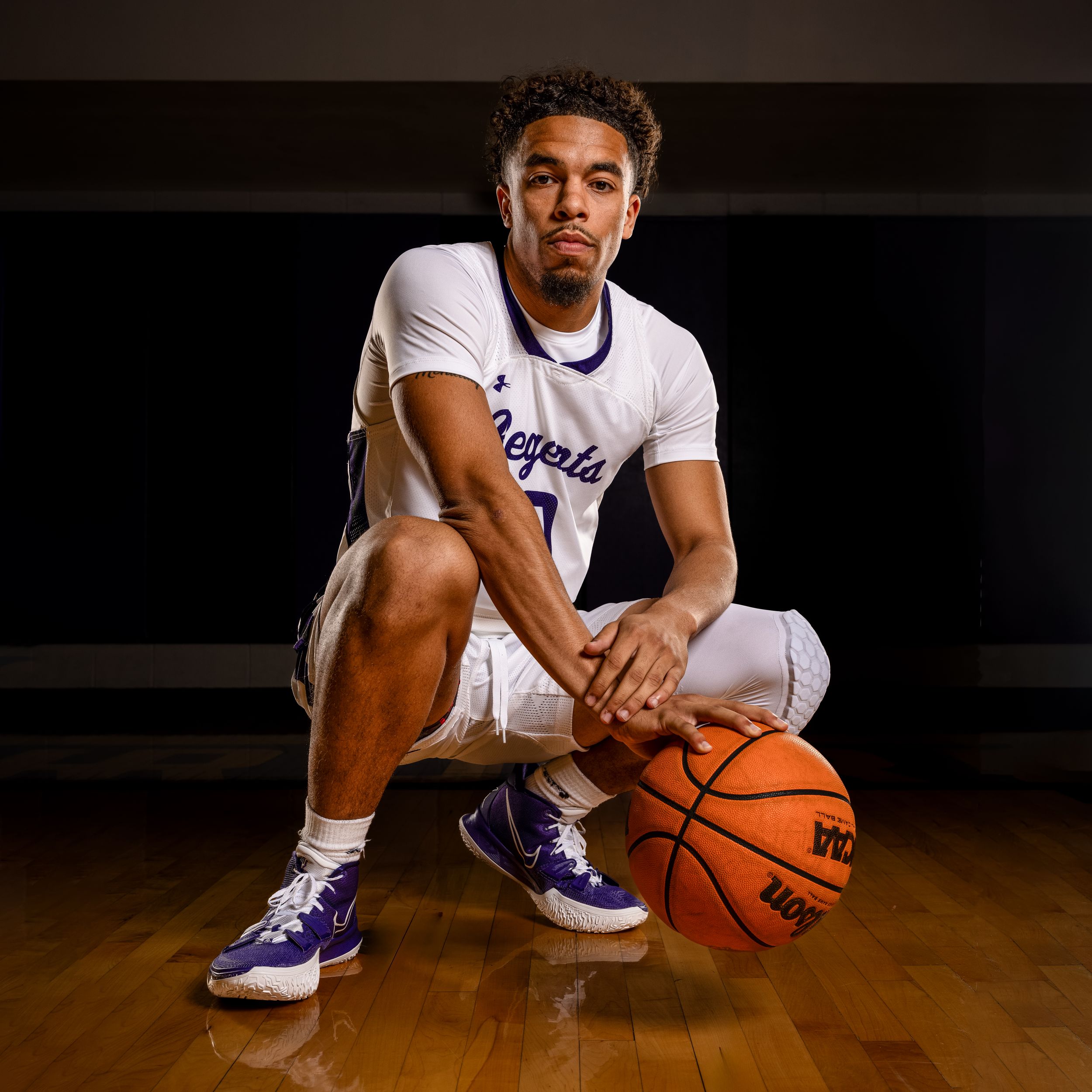Basketball player in white uniform crouches on hardwood court holding a basketball in dramatic lighting.