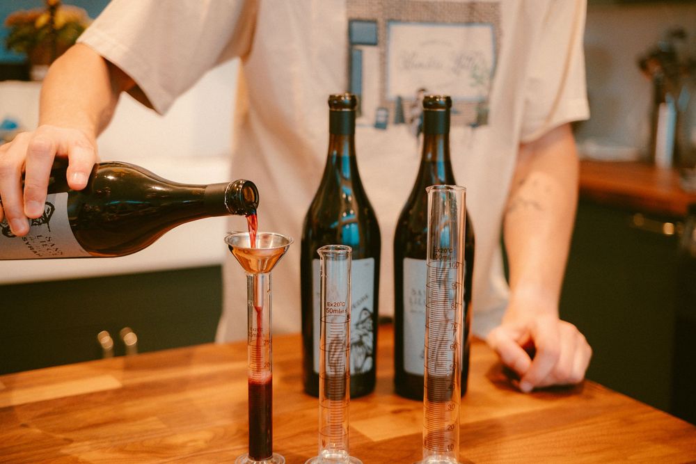 Person pouring red wine through a funnel into a decanter beside two Kelsey wine bottles on a wooden bar.