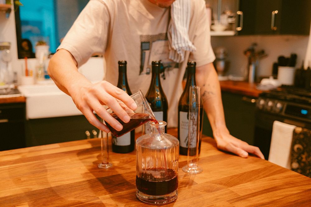 Person decanting red wine into a glass decanter on a wooden counter with Kelsey wine bottles in background.